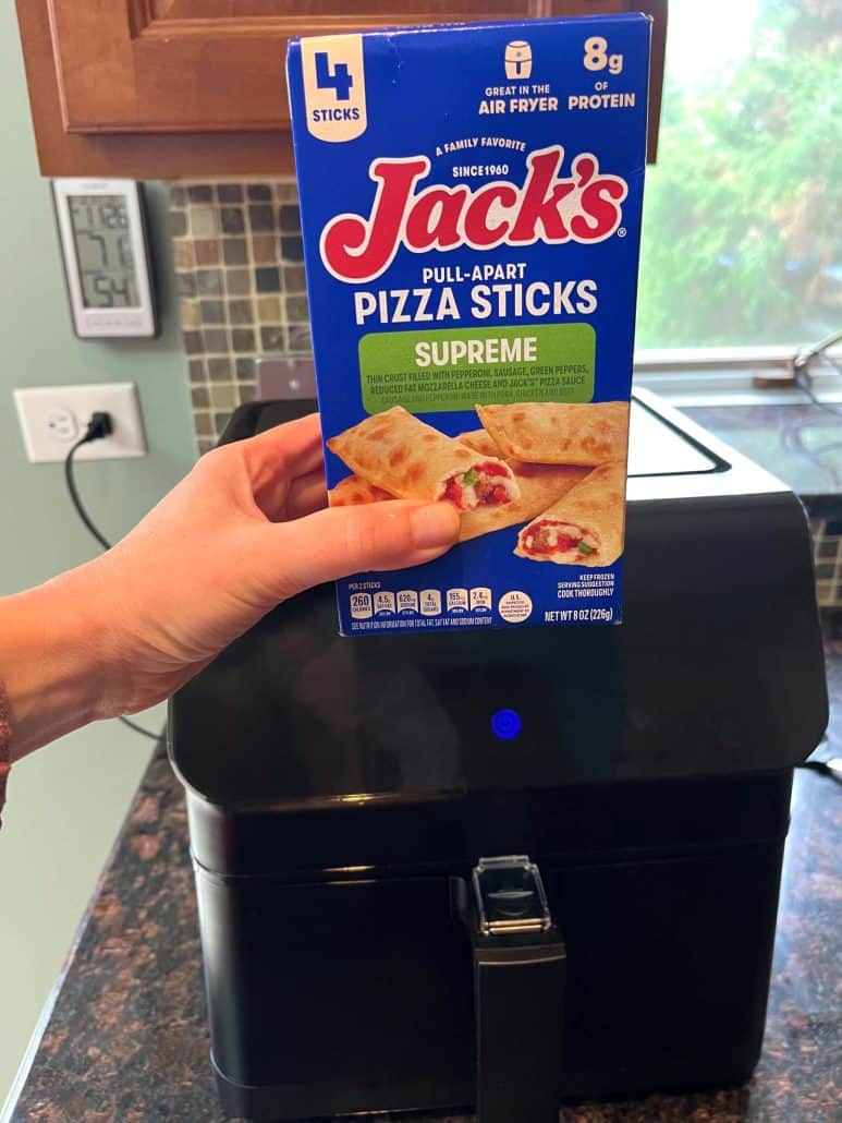 Hand of Melanie Mendelson from melaniecooks.com holding a box of Jack’s Supreme Pizza Sticks in front of an air fryer, ready to prepare a quick and crispy snack. 