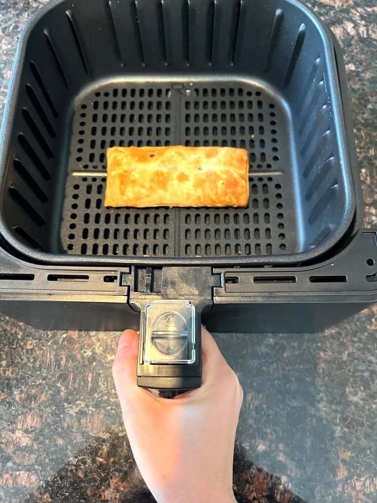 Hand of Melanie Mendelson from melaniecooks.com holding an air fryer basket with a golden-brown pizza stick inside.