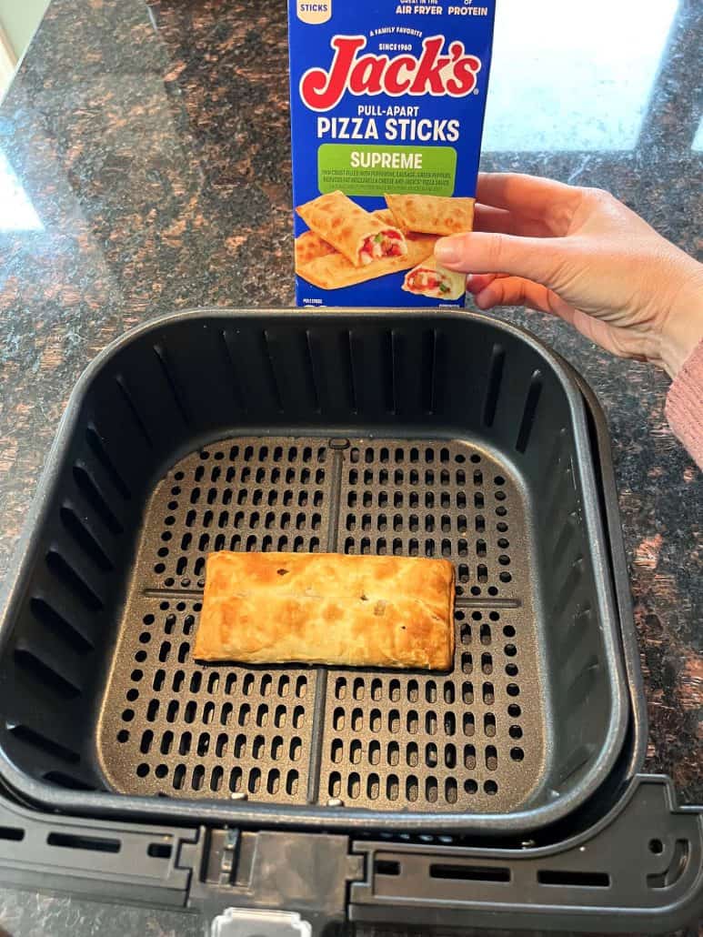 Freshly air-fried pizza stick in the basket, with a hand of Melanie Mendelson from melaniecooks.com holding the Jack’s Supreme Pizza Sticks box in the background.