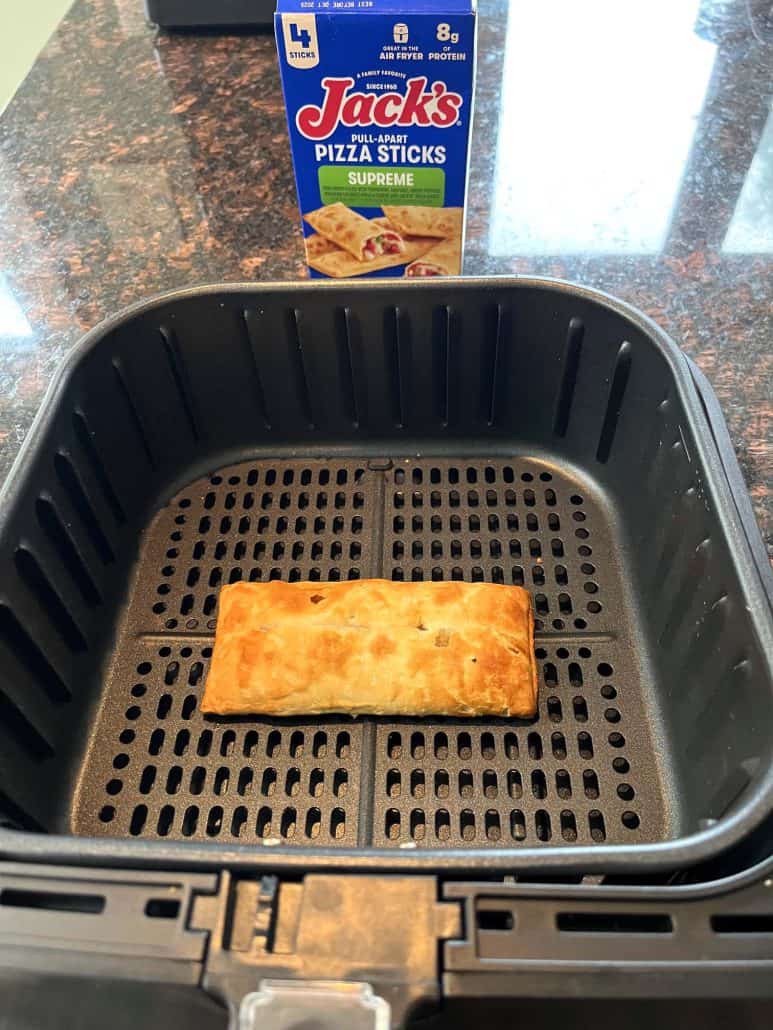 Golden pizza stick in an air fryer basket with the Jack’s Supreme Pizza Sticks box in the background, showcasing the finished product after air frying.