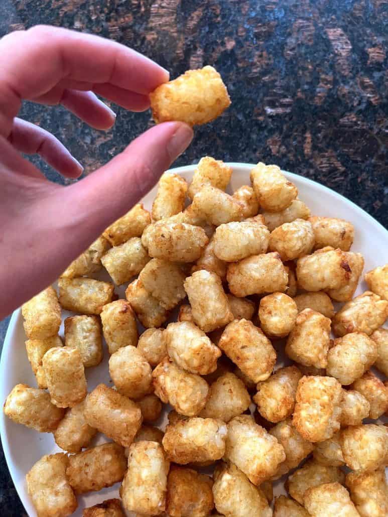 Hand of Melanie Mendelson from melaniecooks.com picking up a crispy Sonic tater tot from a plate filled with golden, perfectly cooked tots.