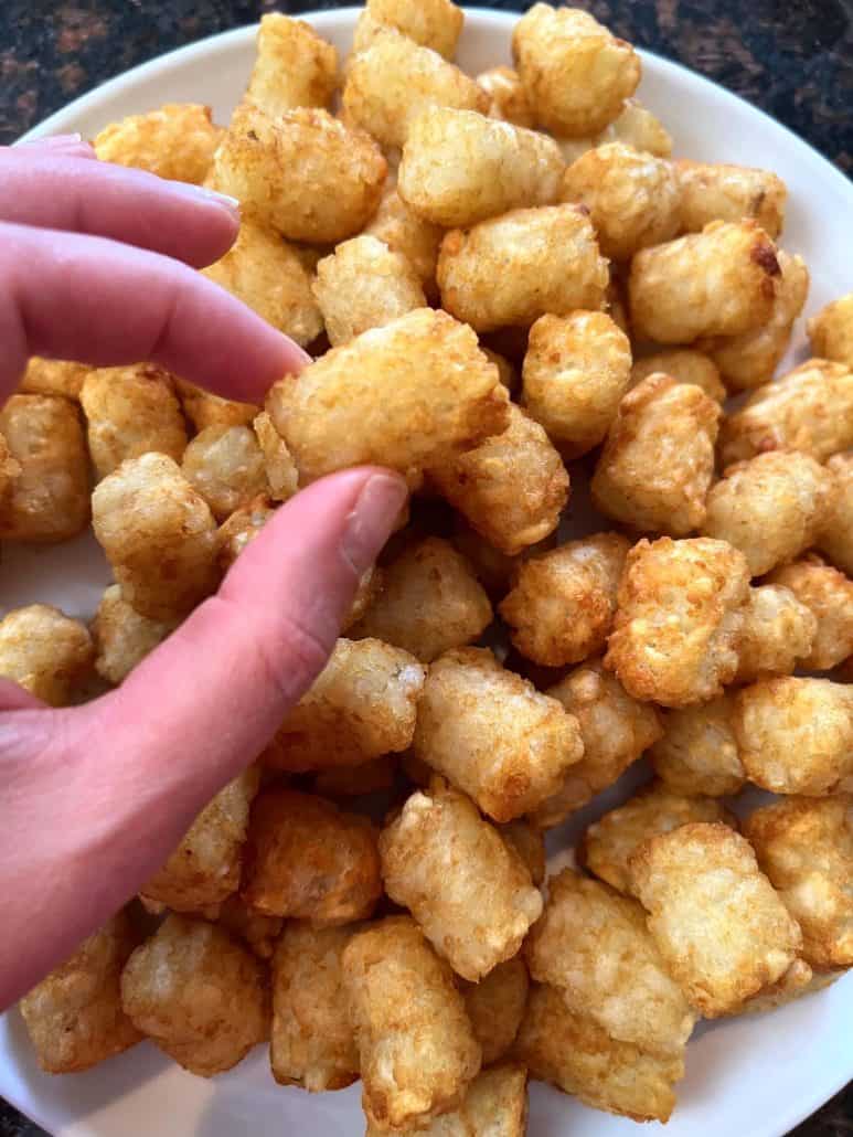 Hand of Melanie Mendelson from melaniecooks.com reaching for a crispy, golden Sonic tater tot from a plate full of freshly cooked tots.