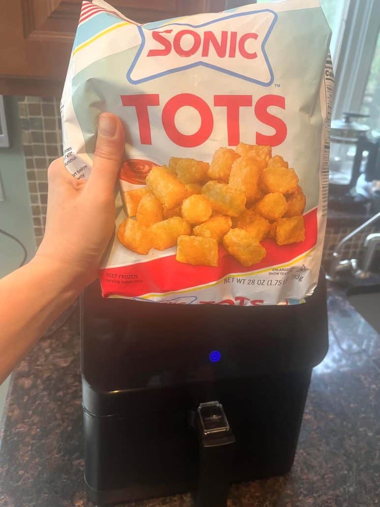 Hand of Melanie Mendelson from melaniecooks.com holding a bag of frozen Sonic tater tots above the air fryer, ready to prepare a crispy and delicious snack.