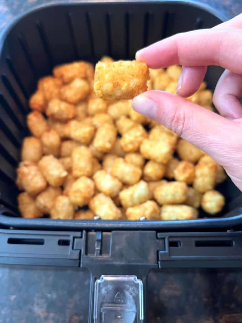 Hand of Melanie Mendelson from melaniecooks.com holding a crispy Sonic tater tot above the air fryer basket filled with golden, perfectly cooked tots.