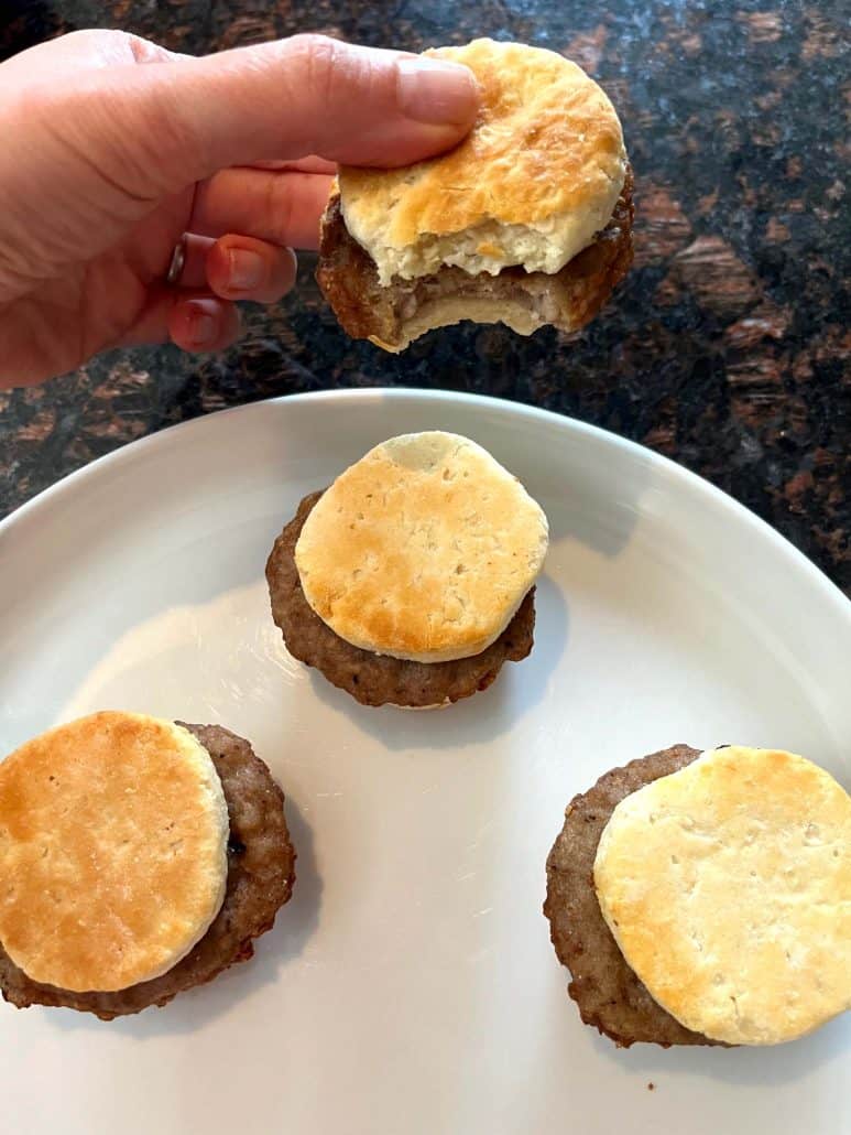 Hand of Melanie Mendelson from melaniecooks.com holding a partially eaten Jimmy Dean Biscuit Sausage Sandwich above a plate with three sandwiches, showcasing the delicious texture and filling.