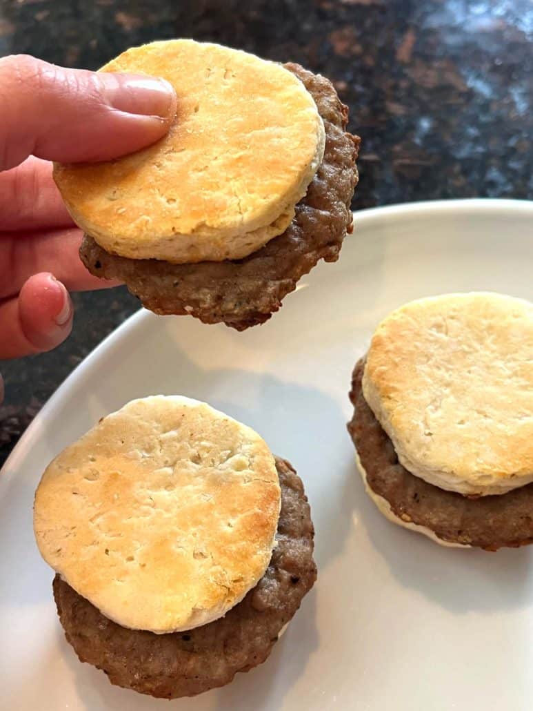 Hand of Melanie Mendelson from melaniecooks.com lifting a cooked Jimmy Dean Biscuit Sausage Sandwich from a plate.