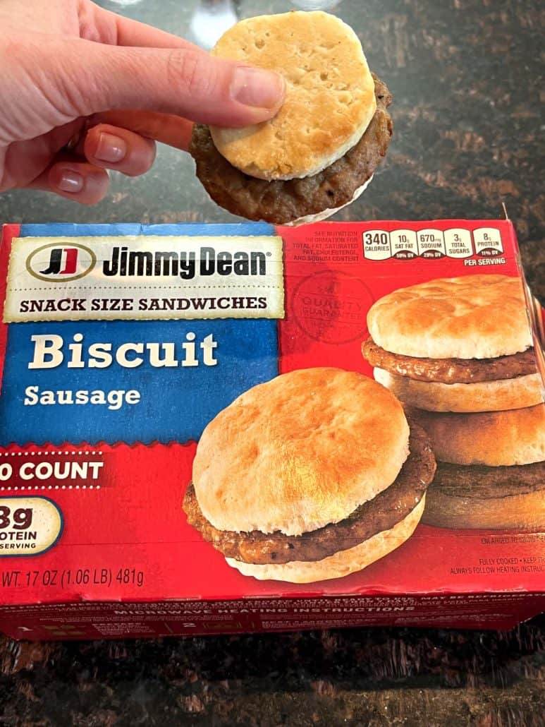 Hand of Melanie Mendelson from melaniecooks.com holding a cooked Jimmy Dean Biscuit Sausage Sandwich above its packaging, showcasing the final result of the air fryer recipe.