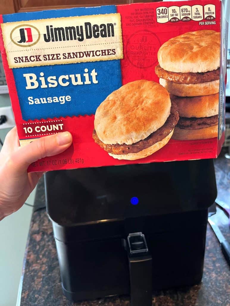 Box of Jimmy Dean Biscuit Sausage Snack Sandwiches held in front of an air fryer, highlighting the preparation step for cooking this easy air fryer recipe.