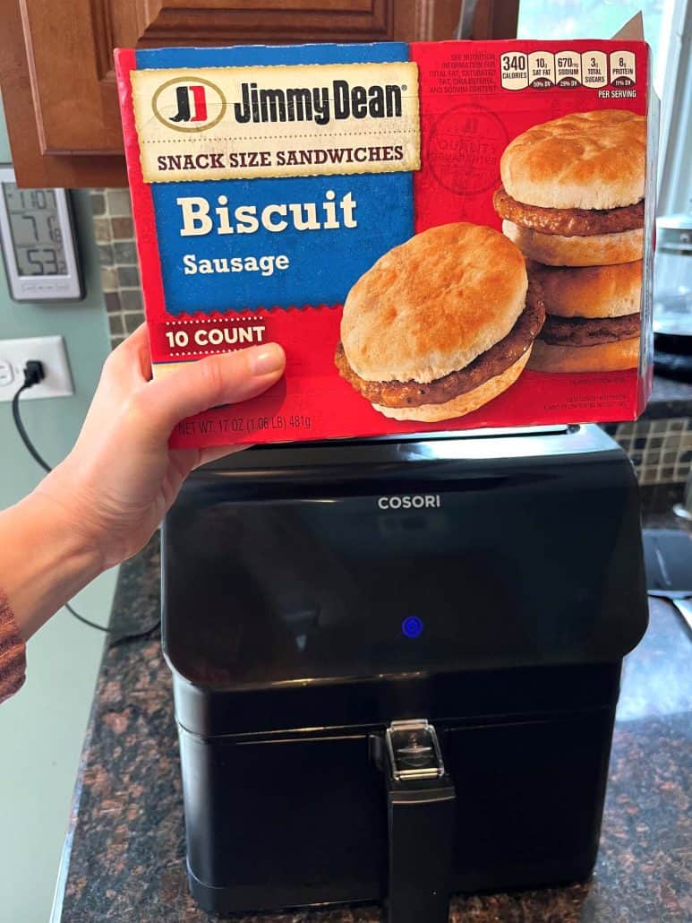 Hand of Melanie Mendelson from melaniecooks.com holding a box of Jimmy Dean Biscuit Sausage Snack Sandwiches in front of an air fryer.