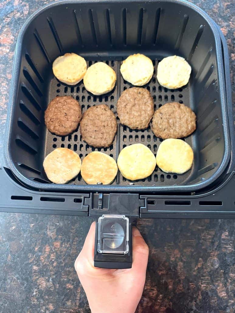 Hand of Melanie Mendelson from melaniecooks.com holding an air fryer basket filled with sausage patties and biscuit halves, prepared for cooking Jimmy Dean Biscuit Sausage Sandwiches.