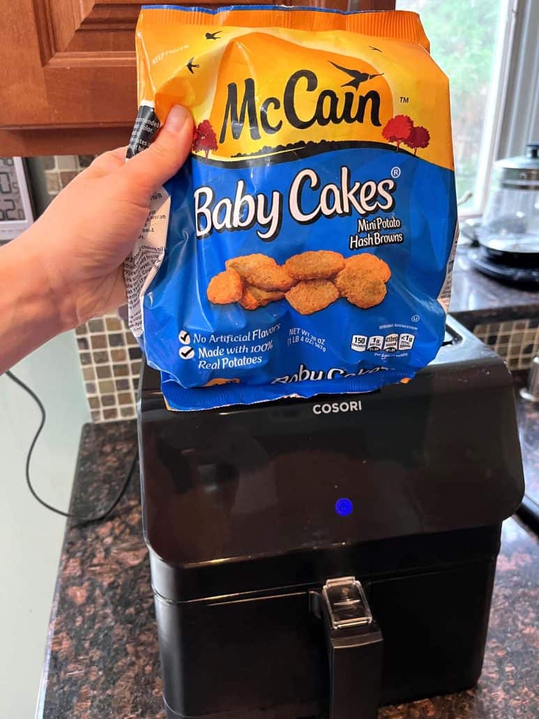 Hand of Melanie Mendelson from melaniecooks.com holding a package of McCain Baby Cakes Hashbrowns above an air fryer, ready to prepare crispy, golden bites.