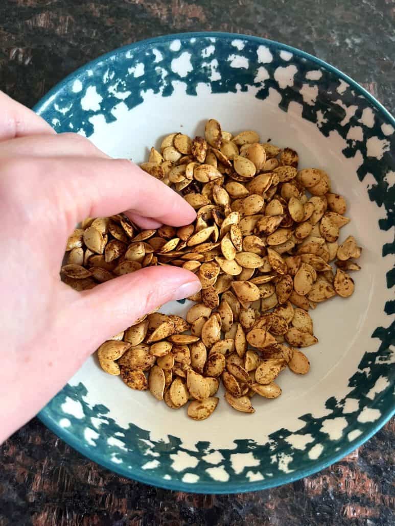 Hand of Melanie Mendelson from melaniecooks.com reaching into a bowl of golden, roasted pumpkin seeds.