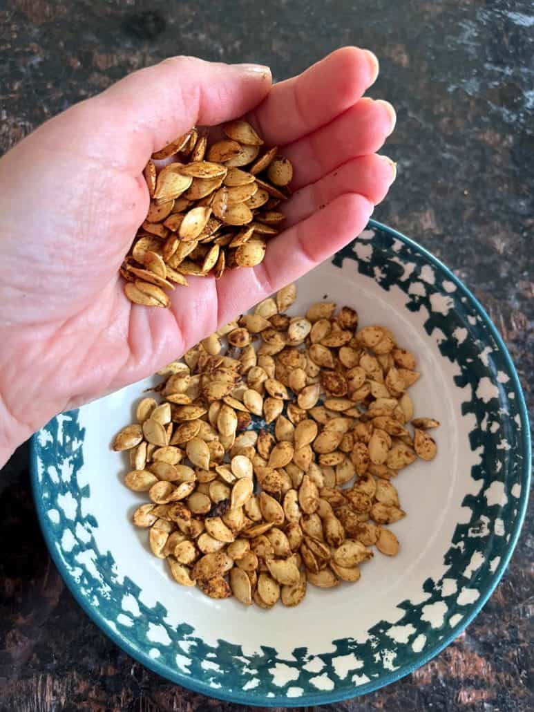 Hand of Melanie Mendelson from melaniecooks.com holding a handful of roasted pumpkin seeds over a bowl, showcasing the golden, crunchy texture of this healthy snack.