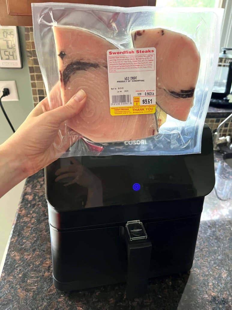 Hand of Melanie Mendelson from melaniecooks.com holding a package of frozen swordfish fillets above an air fryer.