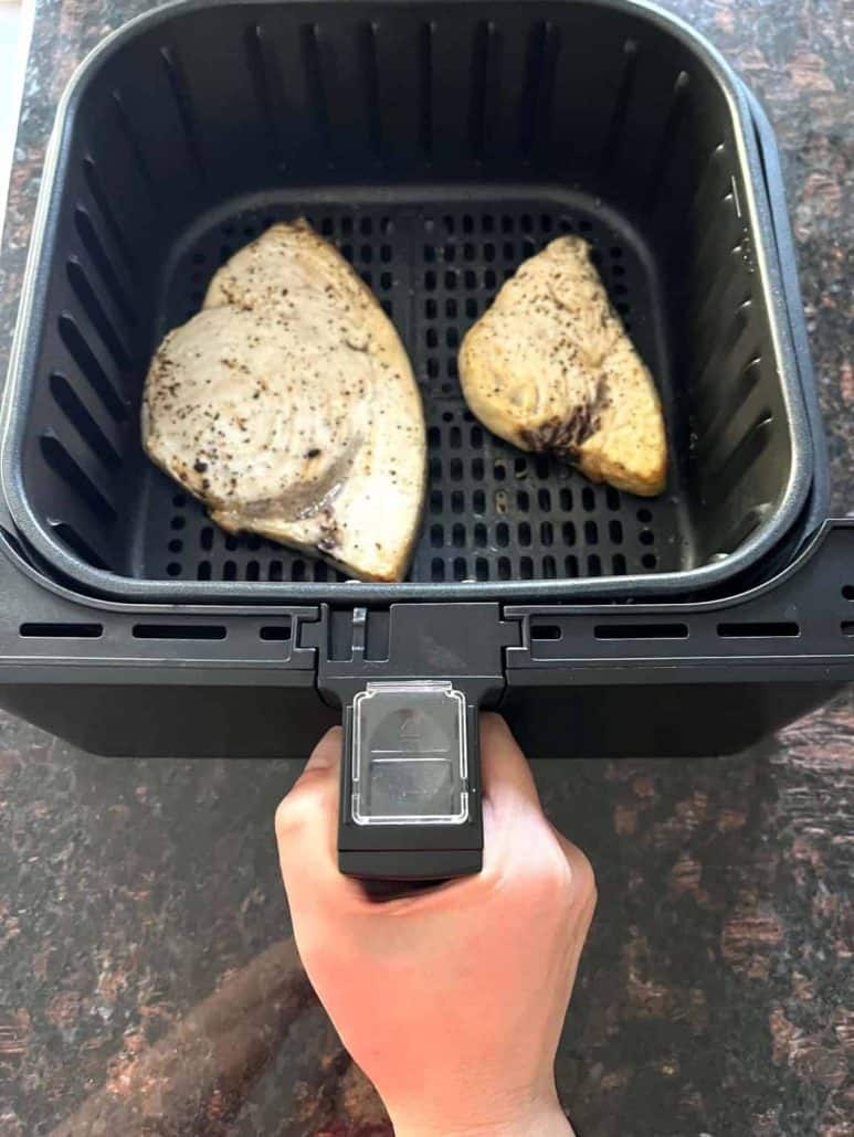 Hand of Melanie Mendelson from melaniecooks.com holding an air fryer basket with two seasoned swordfish fillets inside.