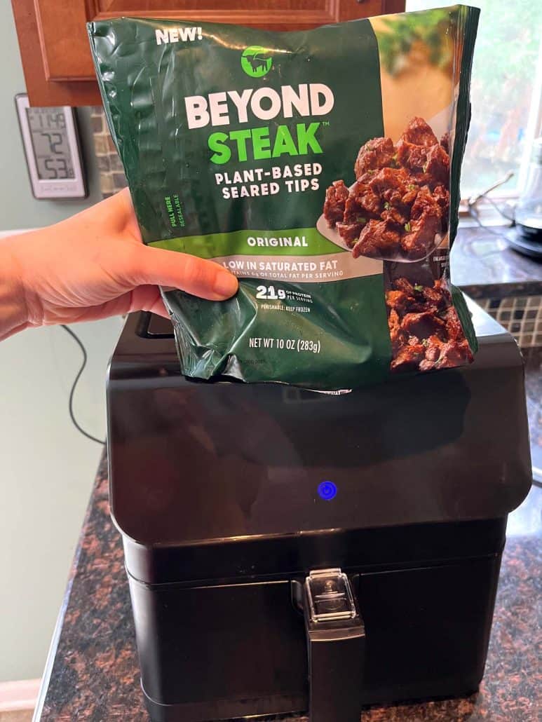 Hand of Melanie Mendelson from melaniecooks.com holding a package of Beyond Steak plant-based tips in front of an air fryer, ready to prepare.