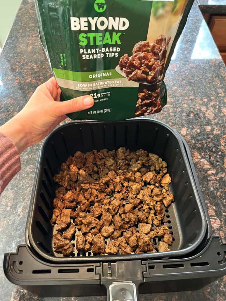 Hand of Melanie Mendelson from melaniecooks.com holding a package of Beyond Steak plant-based tips above an air fryer basket filled with cooked steak tips.