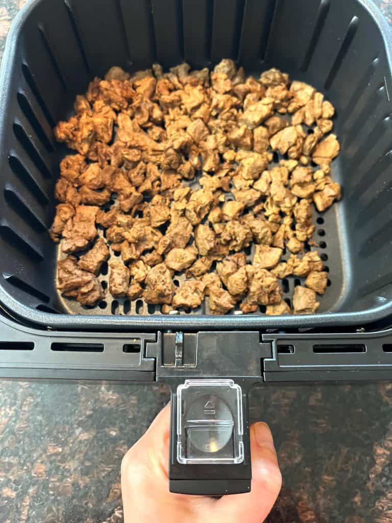 Hand of Melanie Mendelson from melaniecooks.com holding the air fryer basket filled with freshly cooked Beyond Steak Tips, ready to serve as a tasty vegan option.