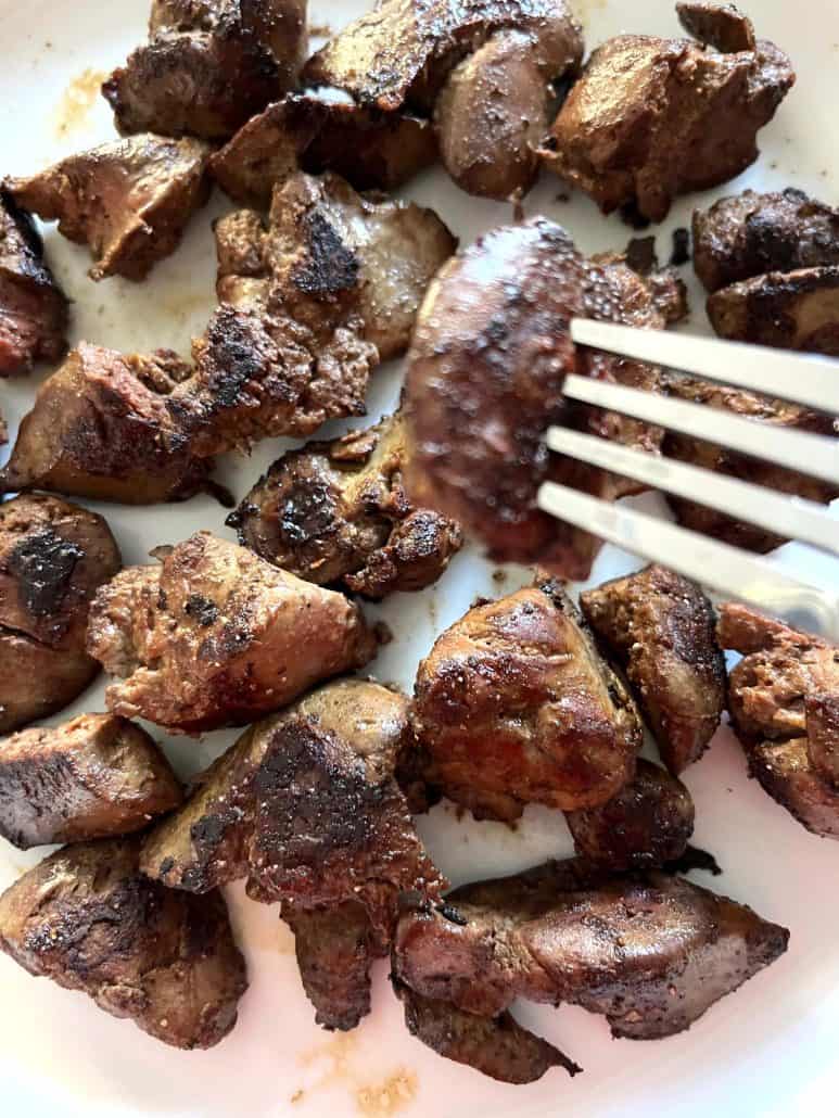 Fork picking up a piece of browned chicken liver from a plate filled with sauteed liver pieces.