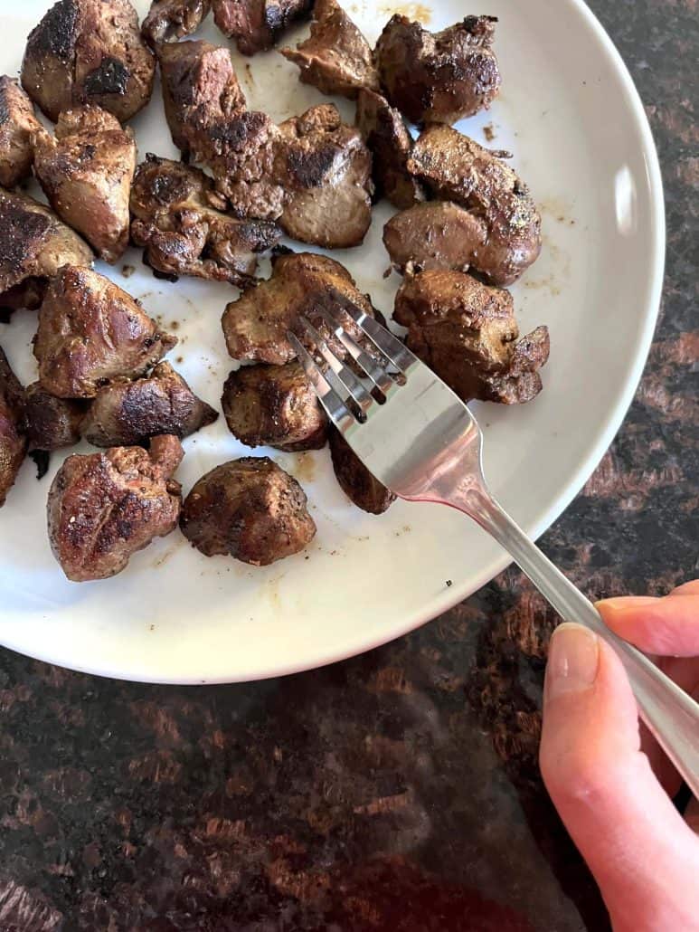 Hand of Melanie Mendelson from melaniecooks.com holding a fork, ready to enjoy freshly cooked, golden-brown chicken livers served on a white plate.