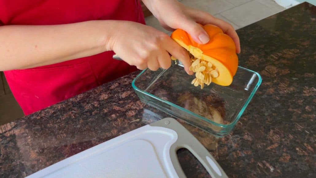 Melanie Mendelson from melaniecooks.com scooping out seeds from a pumpkin half into a glass dish, preparing it for cooking.