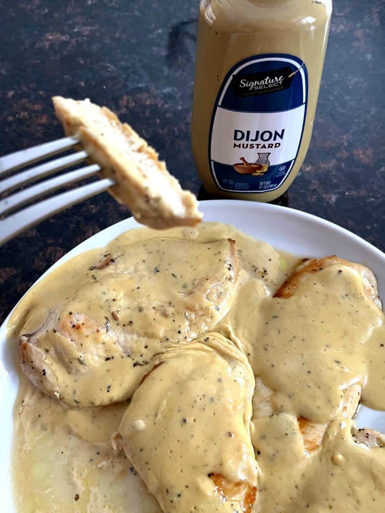 Fork holding a slice of tender Dijon Mustard Chicken above a plate, with a bottle of Dijon mustard in the background.