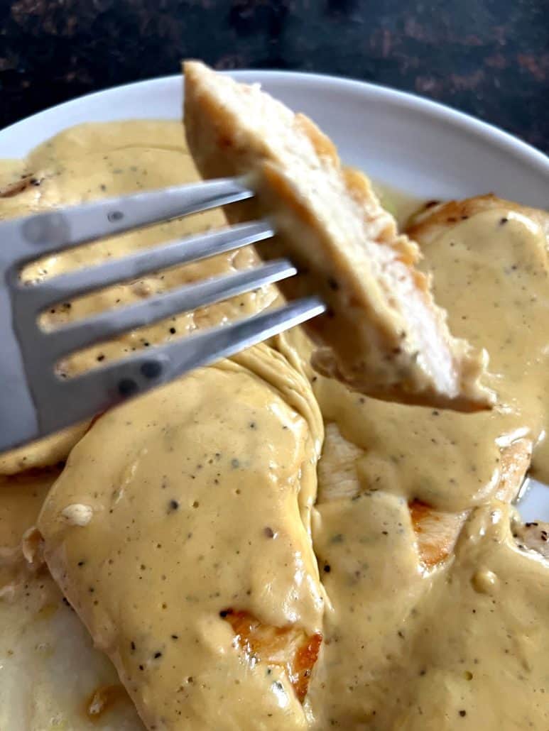 Close-up of a fork holding a slice of Dijon Mustard Chicken, showing the juicy texture and rich creamy sauce on the plate.