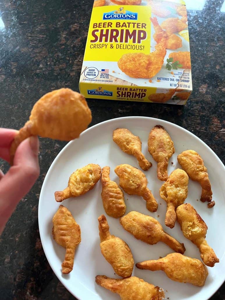 Hand holding a crispy, golden beer-battered shrimp above a white plate filled with more shrimp, with a box of Gorton's Beer Batter Shrimp in the background, highlighting the easy preparation and delicious result.