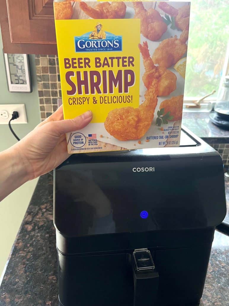 Hand holding a box of Gorton's Beer Batter Shrimp above an air fryer, highlighting the convenience of preparing crispy and delicious shrimp in the air fryer for a quick meal or snack.