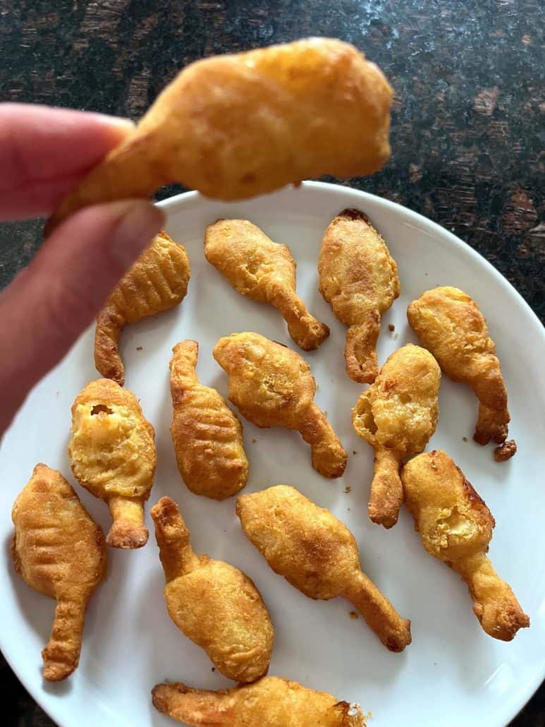 Hand holding a single golden, crispy beer-battered shrimp above a white plate filled with more shrimp, emphasizing their crunchy texture and ready-to-eat appeal