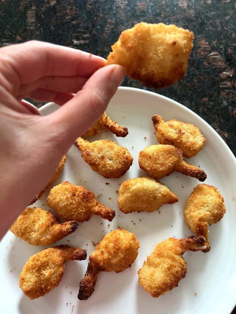 Hand of Melanie Mendelson from melaniecooks.com holding a crispy butterfly shrimp over a plate filled with perfectly cooked shrimp, ready to enjoy.