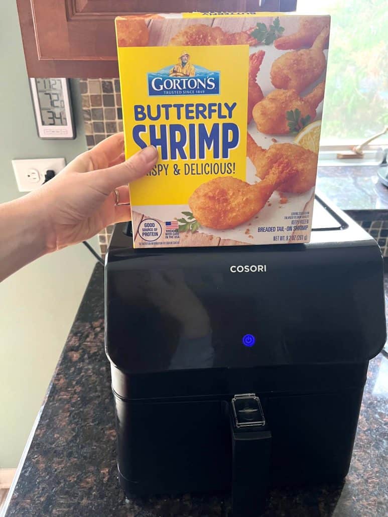 Hand of Melanie Mendelson from melaniecooks.com holding a box of Gorton's Butterfly Shrimp in front of an air fryer, ready for cooking the recipe.