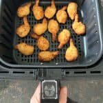 Hand of Melanie Mendelson from melaniecooks.com holding an air fryer basket filled with perfectly cooked butterfly shrimp, ready to serve.