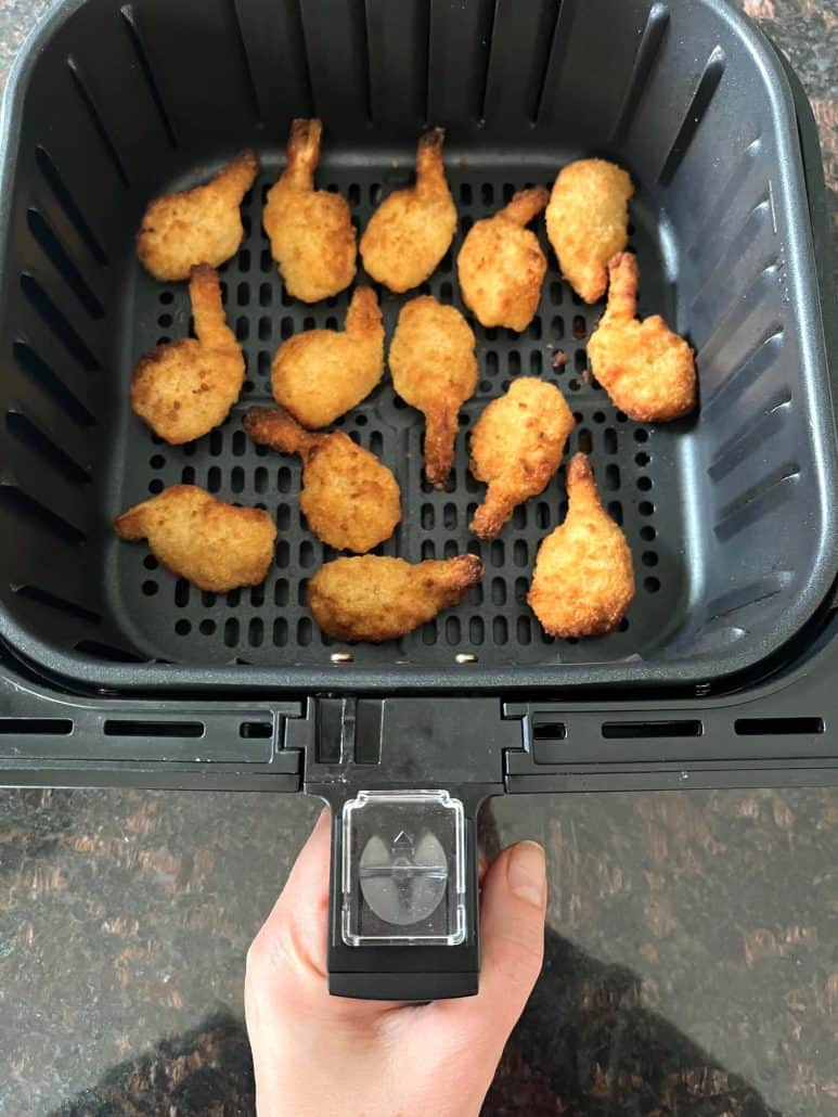 Hand of Melanie Mendelson from melaniecooks.com holding an air fryer basket filled with perfectly cooked butterfly shrimp, ready to serve.