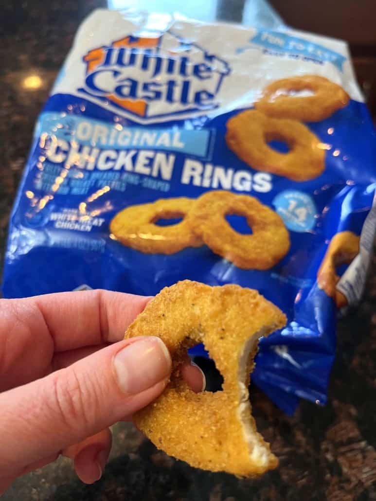 The hand of Melanie Mendelson from melaniecooks.com holding a half-bitten White Castle chicken ring, with the package displayed in the background. Showcasing its crispy golden texture.