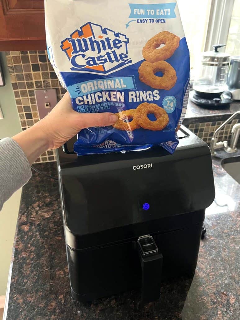 The hand of Melanie Mendelson from melaniecooks.com is holding a package of White Castle Original Chicken Rings above an air fryer.