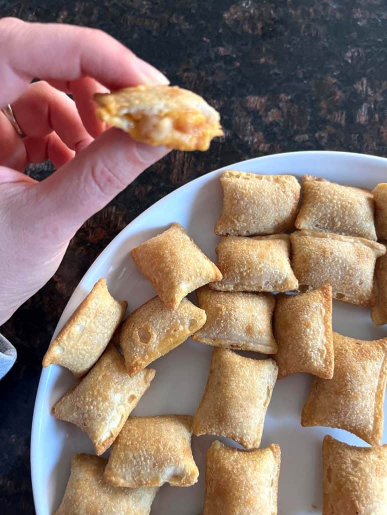 Hand of Melanie Mendelson from melaniecooks.com holding a Totino's Breakfast Snack Bite with a gooey cheesy filling exposed, alongside a plate of golden bites ready to enjoy.