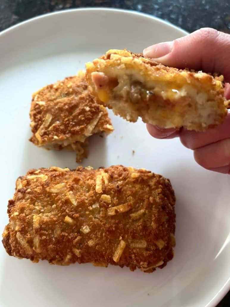 Close-up hand of Melanie Mendelson from melaniecooks.com holding a Jimmy Dean stuffed hashbrown split open to reveal the cheesy filling, with additional crispy hashbrowns on a plate below.