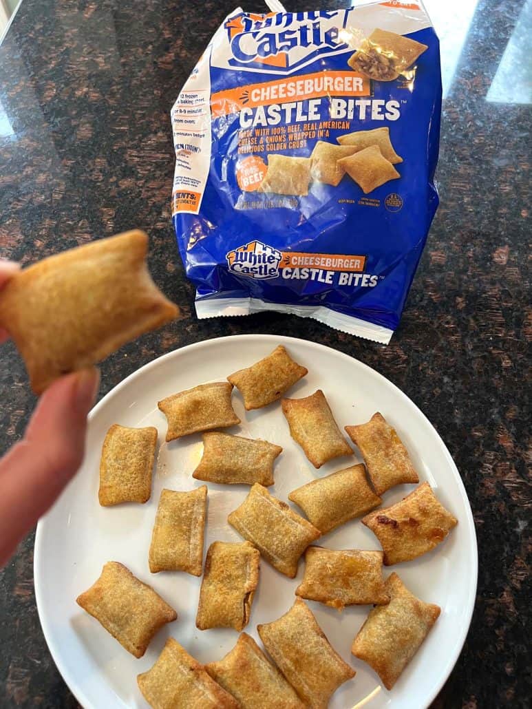 Hand of Melanie Mendelson from MelanieCooks.com holding a crispy, golden-brown White Castle Cheeseburger Bite above a plate of bites, with the branded package in the background, highlighting this easy and delicious snack idea.