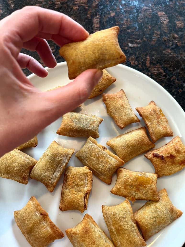 Hand holding a single White Castle Cheeseburger Bite above a white plate filled with more golden-brown bites, emphasizing their crispy texture and convenient, bite-sized design for snacking or sharing.