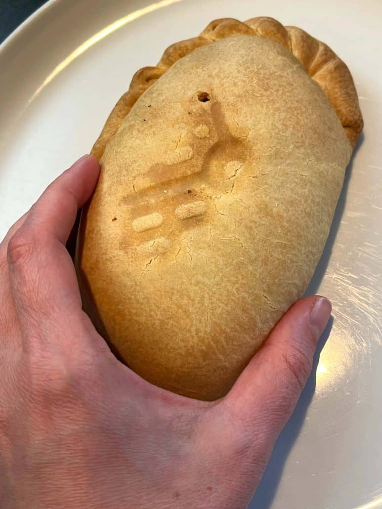 The hand of Melanie Mendelson from melaniecooks.com holding a freshly air-fried pasty on a white plate, showing its crispy, golden crust and ready-to-eat texture.