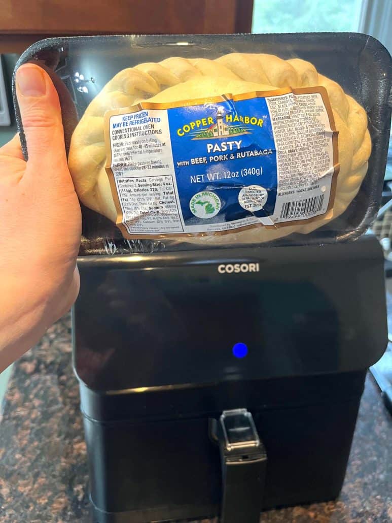 The hand of Melanie Mendelson from melaniecooks.com holding a packaged frozen pasty in front of an air fryer, shows the easy preparation process before cooking.