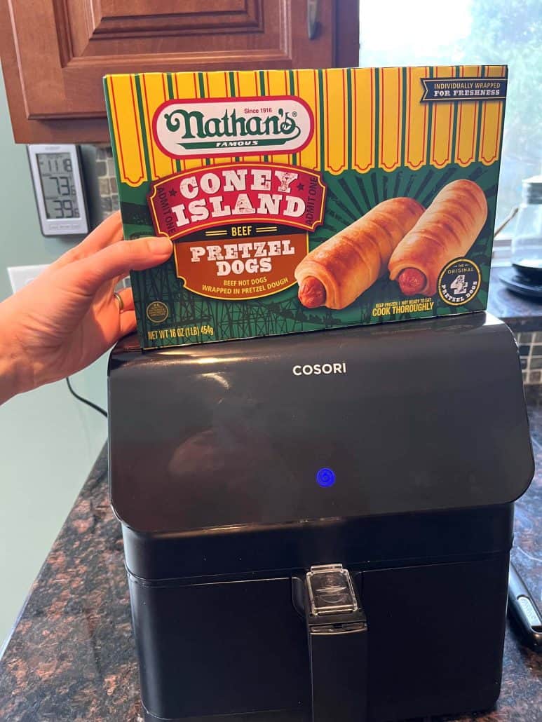 The hand of Melanie Mendelson from melaniecooks.com holding a box of Nathan’s Coney Island frozen pretzel dogs above an air fryer, ready for cooking.