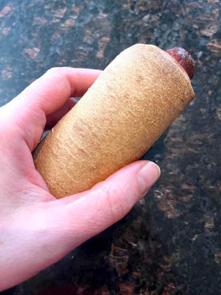 Hand of Melanie Mendelson from melaniecooks.com holding a freshly cooked pretzel dog with a golden-brown crust.