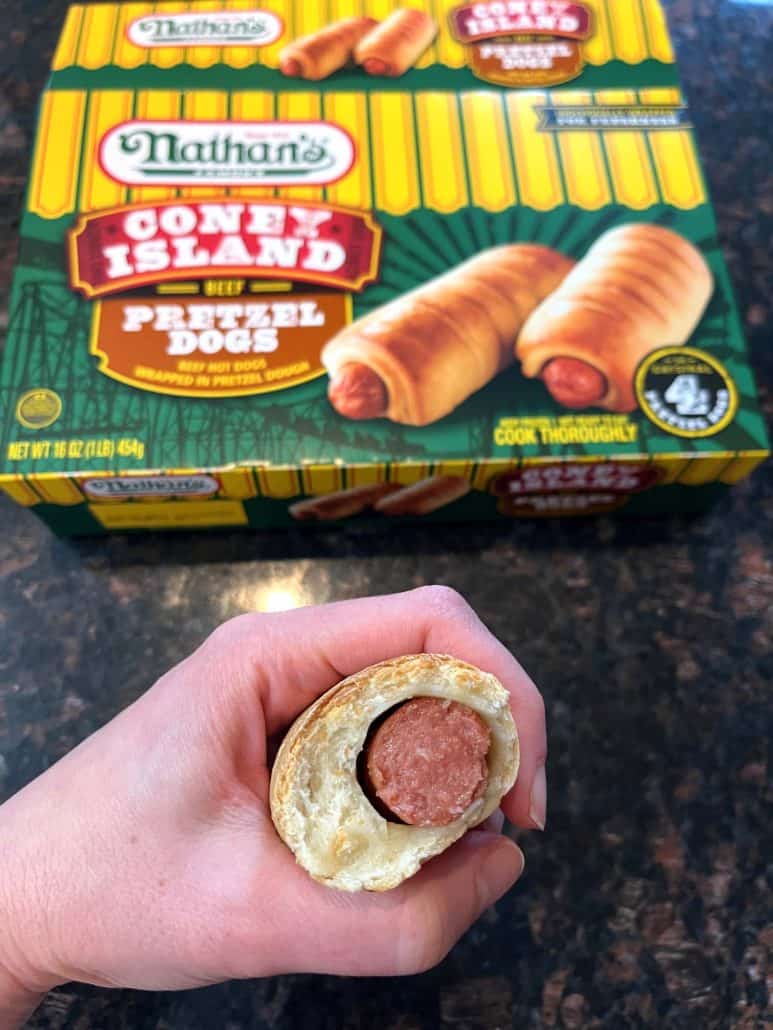 The hand of Melanie Mendelson from melaniecooks.com holding a bitten pretzel dog, revealing the juicy hot dog inside, with a Nathan’s Coney Island frozen pretzel dog box in the background.