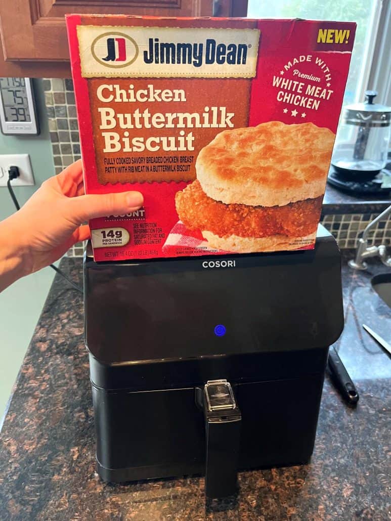 Melanie Mendelson from melaniecooks.com holding a box of Jimmy Dean Chicken Buttermilk Biscuit Sandwiches over an air fryer, ready to cook for a crispy and delicious breakfast.