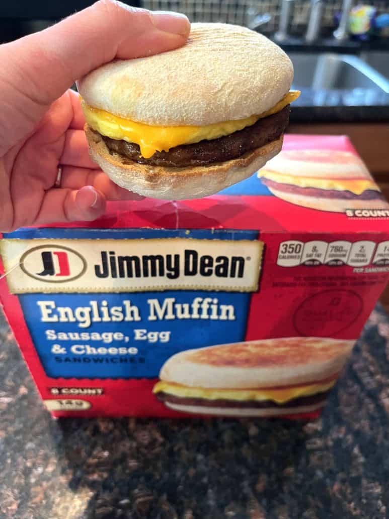 The hand of Melanie Mendelson from melaniecooks.com holding a freshly cooked Jimmy Dean English Muffin Sandwich with sausage, egg, and melted cheese, in front of its original packaging.