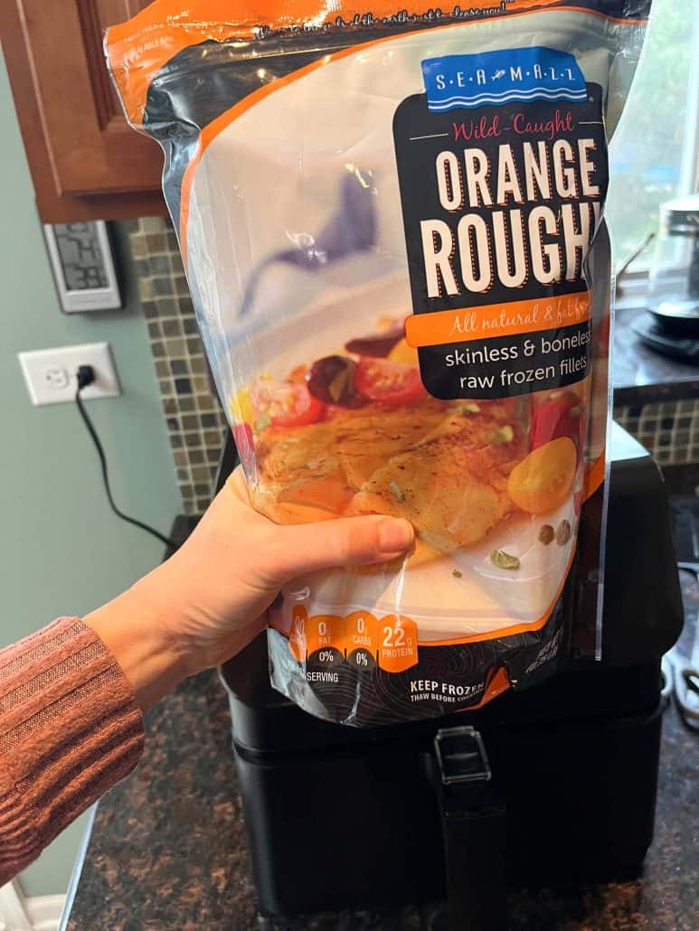 Melanie Mendelson from melaniecooks.com Holding a package of frozen orange roughy fillets in front of an air fryer, ready to cook.