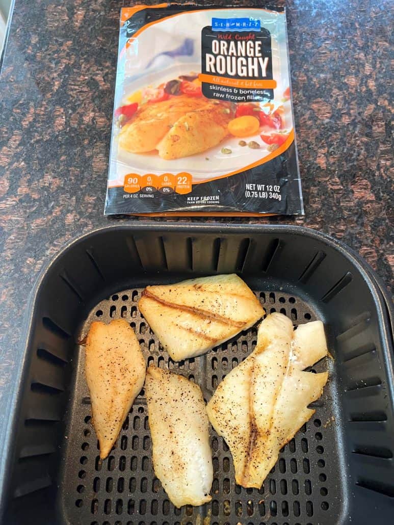 Golden, air-fried orange roughy fillets in the air fryer basket, with the frozen fish package in the background. 