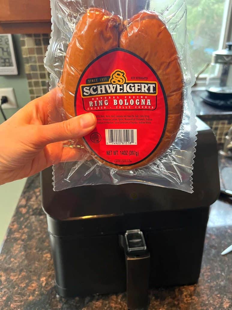 Melanie Mendelson from melaniecooks.com holding a package of ring bologna in front of an air fryer, ready to be sliced and cooked.